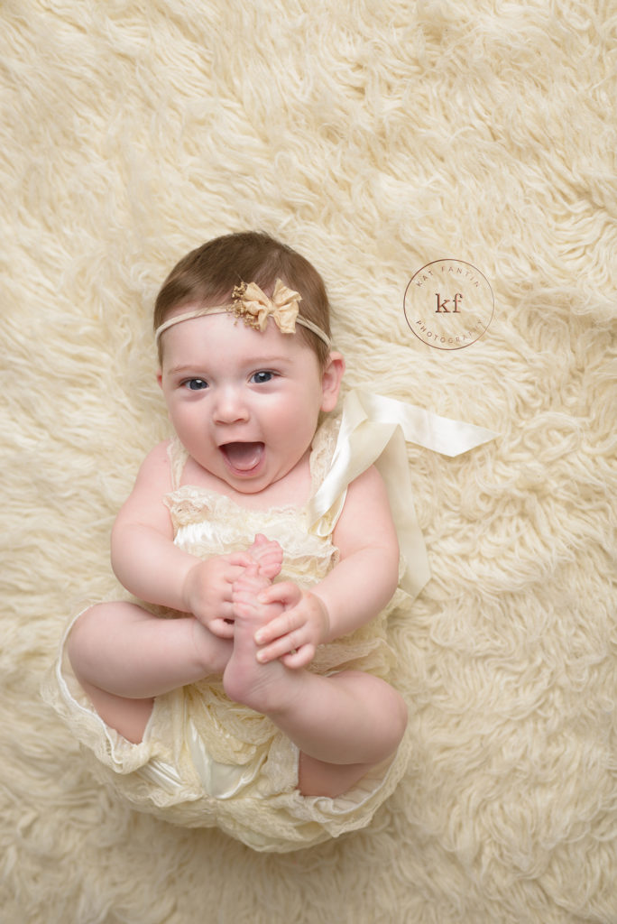 six month old girl from north branch smiles at camera for milestone session with kat fantin with white fur rug and matching romper and headband